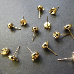 May include: A group of gold-toned earring findings with a round, flat base and a post for attaching earrings. The findings are scattered on a gray surface.