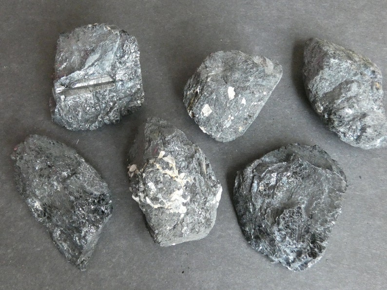 May include: Six black, rough, and irregularly shaped stones with a slightly metallic sheen. The stones have a variety of textures and some have white streaks.