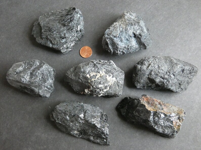 May include: Seven black, rough, and irregularly shaped stones. The stones are various sizes and have a dark, almost metallic sheen. A copper penny is included for scale.