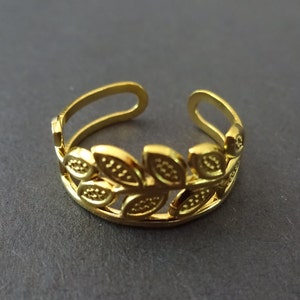 May include: A gold-colored adjustable ring featuring a laurel leaf design. The ring has an open design and is made of a shiny, metallic material. The leaves are detailed with small dots.