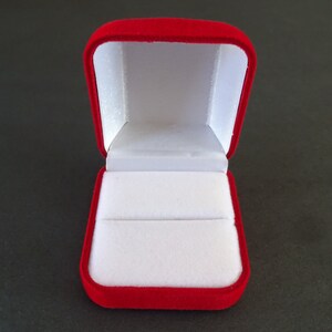 May include: Open red velvet jewelry box with a white interior. The box is rectangular with a hinged lid and a slot to hold a ring or other small item. The interior is lined with white fabric.