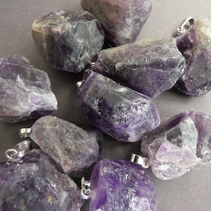 May include: A group of ten raw amethyst crystal pendants with silver bail loops. The crystals are various shades of purple and have a rough, natural texture.