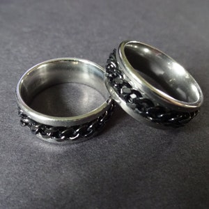 May include: Two silver rings with black chain details. The rings are on a dark gray surface.