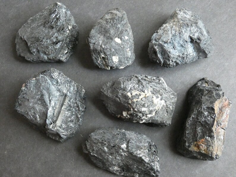 May include: Seven black, rough, and irregularly shaped stones. The stones have a slightly metallic sheen and some have white flecks.