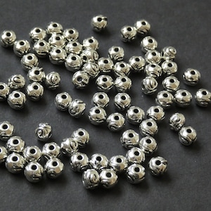 May include: A collection of small, silver-colored metal beads with a textured, patterned surface. Each bead has a hole running through the center. The beads are scattered on a dark surface, suitable for jewelry making or craft projects.