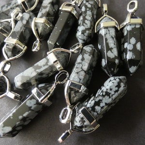 May include: A collection of snowflake obsidian pendants. Each pendant is a hexagonal prism with a silver-colored metal cap and loop. The black stone has white snowflake-like patterns. The pendants are scattered on a dark surface.
