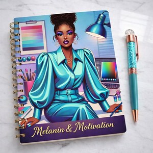 May include: A notebook with a colourful illustration of a woman in a teal outfit, with the text "Melanin & Motivation" on the cover. A teal pen with glitter accents rests beside the notebook. The notebook has gold spiral binding.