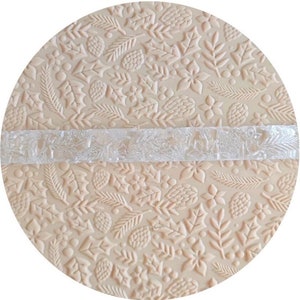 May include: A clear acrylic rolling pin with a raised design of holly leaves, berries, and pine cones. The rolling pin is on top of a textured baking mat with a similar design.
