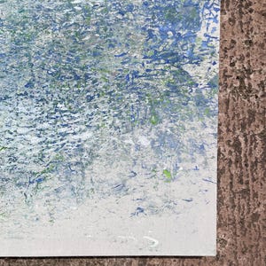 Pode incluir: Pintura abstrata com superf&iacute;cie texturizada. A obra apresenta uma mistura de pinceladas azuis, brancas e verdes, criando um efeito din&acirc;mico e em camadas. A pintura est&aacute; sobre uma tela branca, com uma superf&iacute;cie marrom ao fundo.