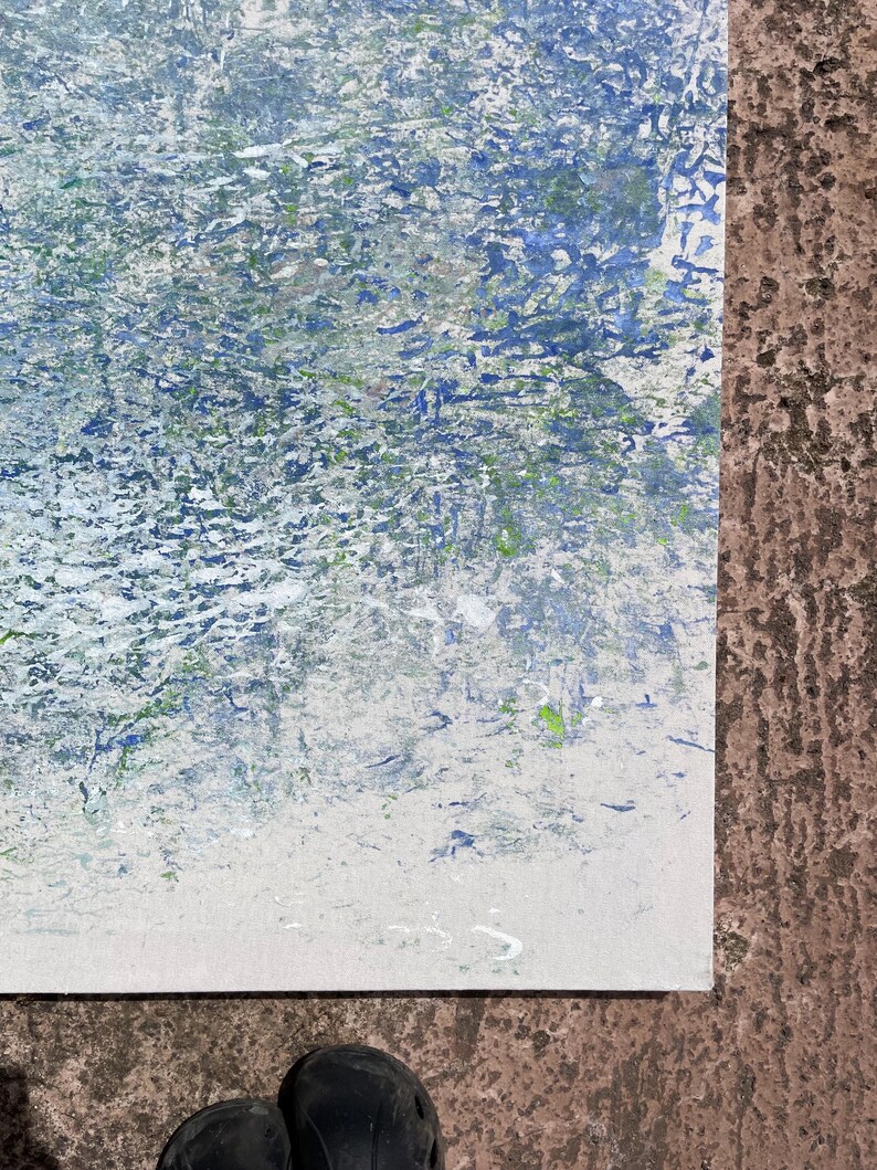 Pode incluir: Pintura abstrata com superf&iacute;cie texturizada. A obra apresenta uma mistura de pinceladas azuis, brancas e verdes, criando um efeito din&acirc;mico e em camadas. A pintura est&aacute; sobre uma tela branca, com uma superf&iacute;cie marrom ao fundo.