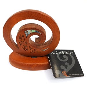 May include: A wooden sculpture with a spiral design, featuring intricate carvings and inlaid iridescent material. The sculpture sits on a round base. A small black tag with the word "Whakairo" is next to the sculpture.