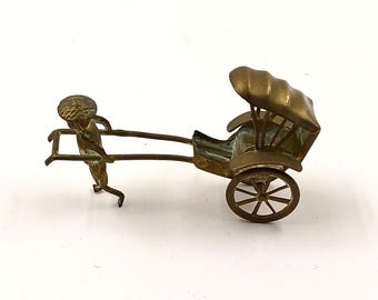 May include: A small brass figurine of a person pulling a rickshaw. The rickshaw has a canopy and a single wheel. The figurine is detailed and has a realistic appearance.