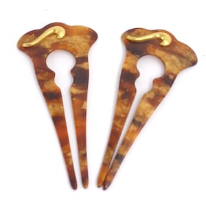 Peut inclure: Deux épingles à cheveux décoratives. Chaque épingle présente un motif écaille de tortue dans des tons marron et or, avec un accent doré sur le dessus. Les épingles ont une forme unique avec une découpe centrale et deux dents.