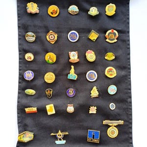 Op de afbeelding: Een verzameling kleurrijke emaille pins tentoongesteld op een zwarte stoffen rol. De pins tonen diverse ontwerpen, waaronder logo's, bezienswaardigheden en emblemen. De pins hebben verschillende vormen en maten, met een verscheidenheid aan kleuren en details.
