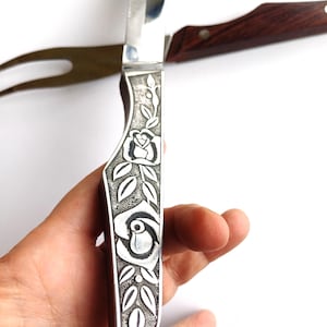 Puede incluir: Un cuchillo para trinchar de color plateado con un diseño floral en el mango. El mango presenta un patrón de rosas y hojas. Un tenedor para trinchar con mango de madera está en el fondo.