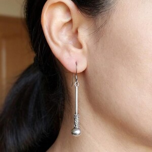 Puede incluir: Un pendiente plateado con un diseño de barra larga y delgada. El pendiente presenta un patrón decorativo cerca de la parte superior y una pequeña bola redonda en la parte inferior. El pendiente está suspendido de un gancho.