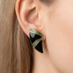 May include: A black and green geometric earring with gold-coloured trim is shown on an ear. The earring has a triangular shape with a black enamel base and green accents. The earring is a clip-on style.