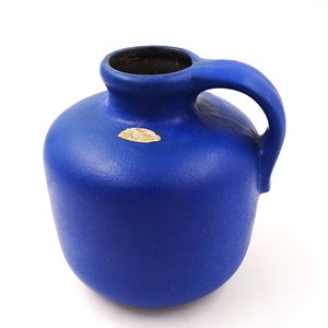 Peut inclure: Un vase en céramique bleu avec une anse. Le vase a un corps arrondi et une surface texturée.