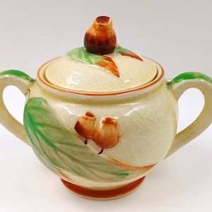 May include: A cream-coloured ceramic sugar bowl with a lid. The bowl has a green and brown floral design with a brown knob on the lid.