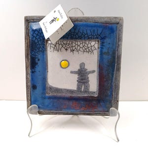 Puede incluir: Un azulejo de cerámica cuadrado con un borde azul y blanco y un centro blanco que presenta una figura de inukshuk gris con un sol amarillo en el fondo. El azulejo está sobre un soporte de plástico transparente. El azulejo está firmado "Carl Veldhuis" y "Hand Crafted".