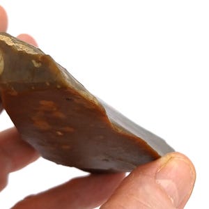 May include: A brown and tan stone tool with a sharp edge, held in a hand. The stone has a smooth, polished appearance with colour and texture variations. The tool is against a white background.