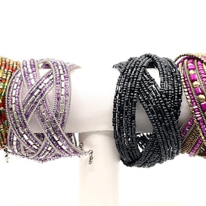 May include: Four beaded bracelets in various styles and colors are displayed. The bracelets include gold, red, and green beads, a lavender woven design, black beads, and a pink and gold design.