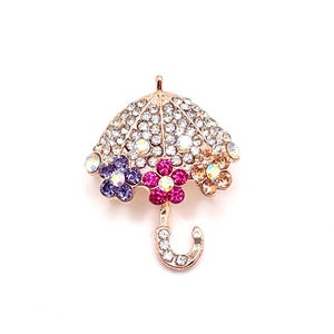 May include: A gold-toned brooch shaped like an umbrella with clear, iridescent, and colored rhinestones. The umbrella has three flowers on the top, one purple, one pink, and one orange.