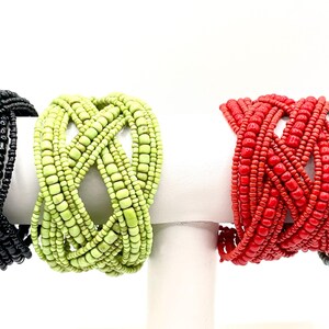 May include: Four beaded bracelets in black, green, red, and a mix of orange, white, and blue. Each bracelet is made of small, evenly sized beads woven together in a crisscross pattern. The bracelets are displayed on white cylindrical stands.