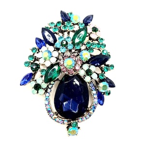 May include: A large, ornate brooch featuring a cluster of colorful gemstones. The brooch has a teardrop-shaped, dark blue center stone, surrounded by smaller blue, green, and iridescent crystals. The design resembles a floral arrangement.