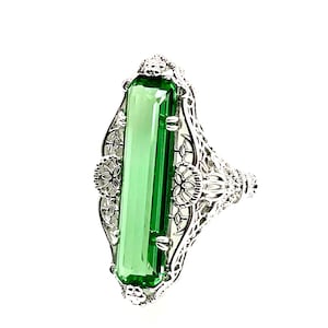 May include: A silver-toned ring featuring a long, rectangular, emerald-green faceted gemstone set vertically. The metal band displays intricate, openwork filigree detailing along the sides, with small floral accents at the top and bottom.