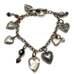 May include: A silver-toned charm bracelet featuring a toggle clasp and a variety of heart-shaped charms. The charms include plain, embossed, and floral designs, interspersed with dark faceted beads and copper-colored jump rings on a chain.