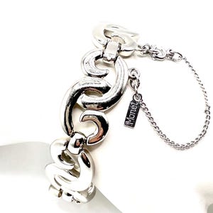 Könnte beinhalten: Ein silberfarbenes Metallgliederarmband mit geschwungenen, ineinandergreifenden Segmenten. Eine feine Sicherheitskette ist am Armband befestigt, das zudem einen kleinen rechteckigen Metallanhänger mit der eingravierten Marke Monet enthält.