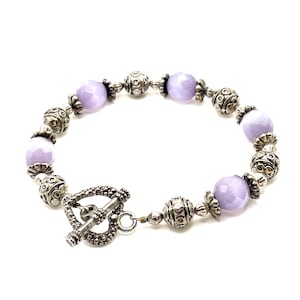 Puede incluir: Una pulsera de cuentas con cuentas facetadas de color lavanda y cuentas ornamentales de metal plateado. La pulsera tiene un cierre de palanca. La joyería se muestra sobre un fondo blanco.