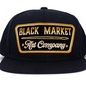 Op de afbeelding: Zwarte baseballpet met platte klep, met een rechthoekige patch. De patch toont "BLACK MARKET Art Company" in goud en wit, met een penseelafbeelding. De pet is zwart.