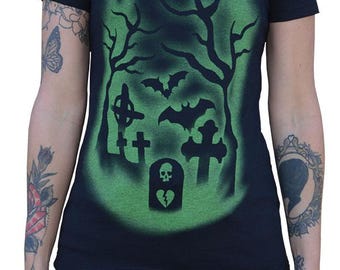 May include: Black t-shirt with a green graveyard design. The graphic includes silhouettes of trees, bats, crosses, and a tombstone with a skull and broken heart. The shirt has short sleeves and a crew neck.