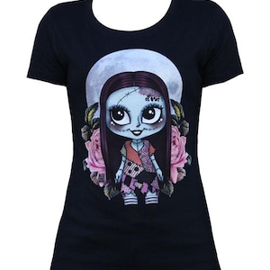 Pode incluir: Camiseta preta com uma ilustra&ccedil;&atilde;o de desenho animado de uma boneca. A boneca tem pele azul, cabelo longo e escuro e um vestido de retalhos. O design inclui uma lua cheia e rosas cor-de-rosa, criando uma est&eacute;tica caprichosa e g&oacute;tica. Uma camiseta gr&aacute;fica &uacute;nica.