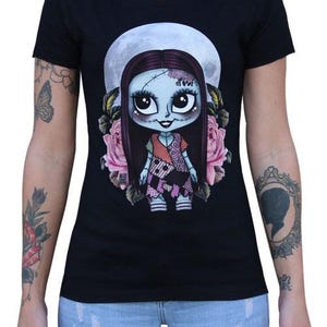 Pode incluir: Camiseta preta com um desenho de boneca de desenho animado, uma lua cheia e rosas cor-de-rosa. A boneca tem pele azul, cabelo longo e escuro e um vestido de retalhos. Uma camiseta gr&aacute;fica &uacute;nica.