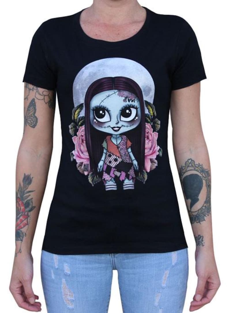 Pode incluir: Camiseta preta com um desenho de boneca de desenho animado, uma lua cheia e rosas cor-de-rosa. A boneca tem pele azul, cabelo longo e escuro e um vestido de retalhos. Uma camiseta gr&aacute;fica &uacute;nica.