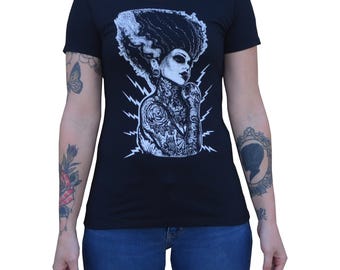May include: Black crew neck t-shirt featuring a white graphic of a woman with elaborate hair and tattoos, set against a backdrop of lightning bolts. The design is centered on the front of the shirt.
