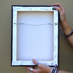 May include: The back of a canvas painting with a black frame. The canvas is stretched over a wooden frame and secured with staples. The painting is titled "La Mujer" by JR Linton. The website "loveheartartcompany.com" is printed on the frame.
