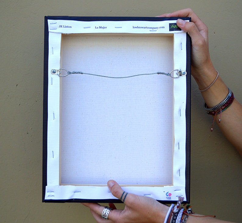 May include: The back of a canvas painting with a black frame. The canvas is stretched over a wooden frame and secured with staples. The painting is titled "La Mujer" by JR Linton. The website "loveheartartcompany.com" is printed on the frame.