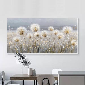 Puede incluir: Una obra de arte rectangular en lienzo que representa un campo de dientes de león. Los dientes de león tienen cabezas de semillas blancas y esponjosas con tallos dorados, sobre un fondo gris y beige apagado. La obra está enmarcada en blanco.
