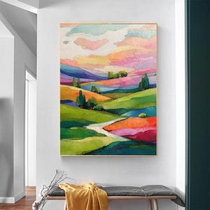 May include: An abstract landscape painting with a gold frame. The artwork features rolling hills in shades of green, orange, purple, and blue, under a sky of pink, yellow, and white. Trees and a winding path add detail to the colorful scene.