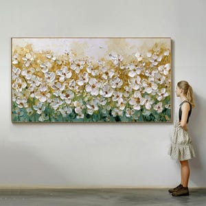 Op de afbeelding: Een groot rechthoekig olieverfschilderij met een veld witte bloemen met getextureerde penseelstreken. De achtergrond gaat over van geel naar groen, omlijst in een goudkleurige houten lijst. Het kunstwerk wordt op een witte muur getoond.