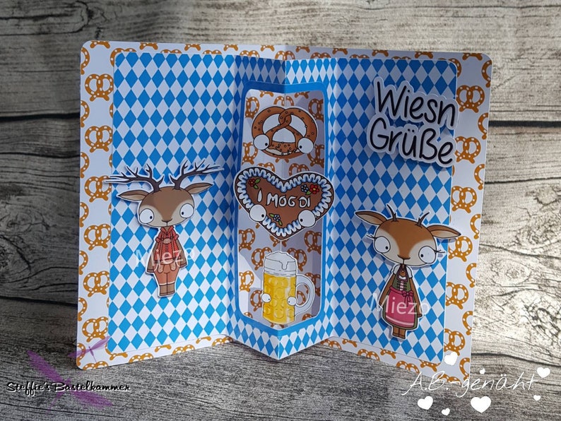 Puede incluir: Una tarjeta a cuadros azules y blancos con un dise&ntilde;o emergente que presenta dos personajes de ciervos, un pretzel, una galleta de jengibre en forma de coraz&oacute;n y una jarra de cerveza. La tarjeta dice "Wiesn Gr&uuml;&szlig;e" e "I mog di".