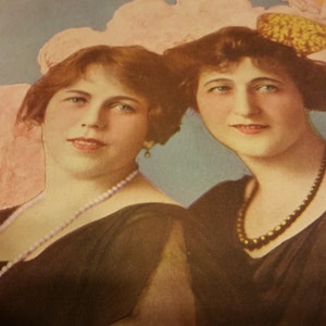 May include: Two women wearing black dresses with pearl and bead necklaces. The woman on the left has short brown hair and the woman on the right has long brown hair with a yellow flower in her hair. The background is a light blue with pink and white flowers.