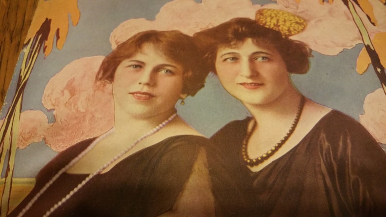 May include: Two women wearing black dresses with pearl and bead necklaces. The woman on the left has short brown hair and the woman on the right has long brown hair with a yellow flower in her hair. The background is a light blue with pink and white flowers.