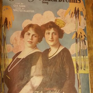 May include: A vintage sheet music cover for the song "My Isle of Golden Dreams" by Gus Kahn and Walter Blaufuss. The cover features a colorful illustration of two women in black dresses, with a palm tree and a sunset in the background. The text "Featured by Holt and Rosedale in "Hello Alexander"" is printed at the bottom.