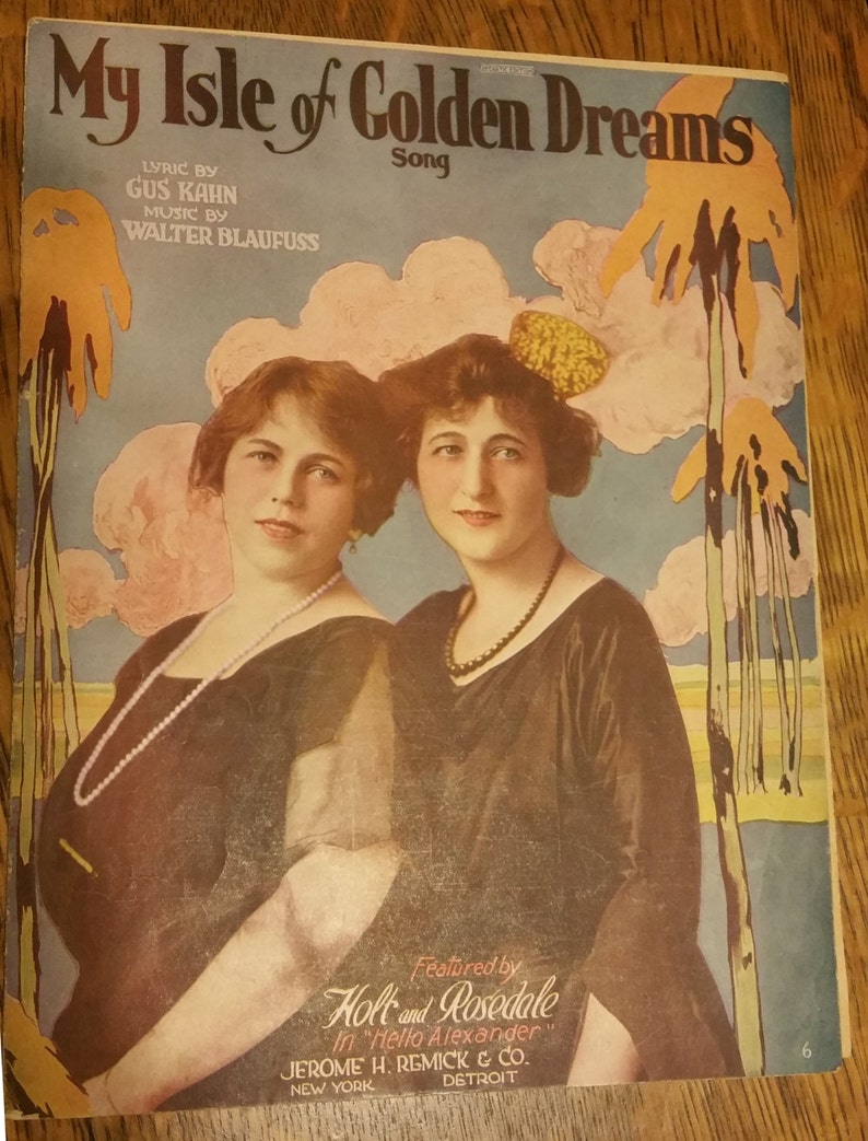 May include: A vintage sheet music cover for the song "My Isle of Golden Dreams" by Gus Kahn and Walter Blaufuss. The cover features a colorful illustration of two women in black dresses, with a palm tree and a sunset in the background. The text "Featured by Holt and Rosedale in "Hello Alexander"" is printed at the bottom.