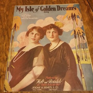 May include: A vintage sheet music cover for the song "My Isle of Golden Dreams" featuring two women in black dresses. The cover features a tropical scene with palm trees and clouds.